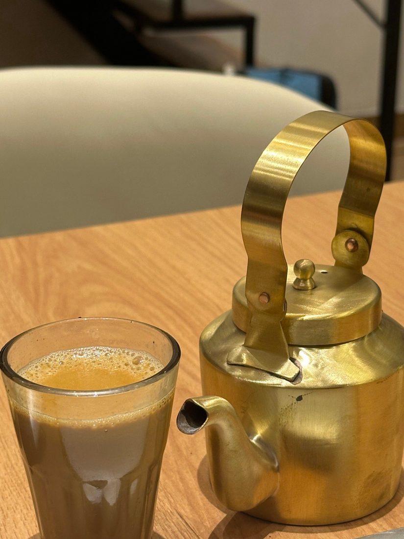 A glass of chai next to a brass kettle