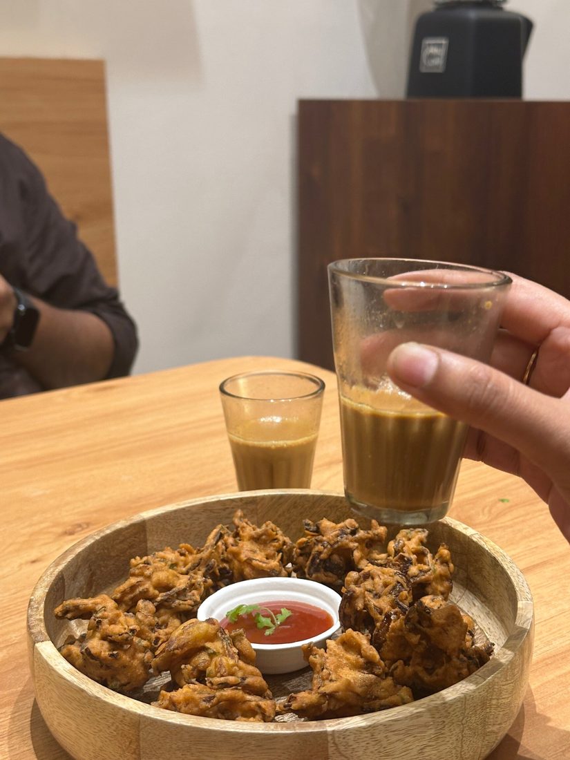 Two glasses of chai raised over a wooden bowl of onion pakoda